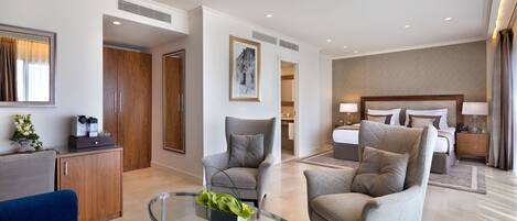 Una habitación de hotel moderna con una mesa de centro de vidrio, dos sillones, un sofá, una cama con cabecero y una mesita de noche con lámparas.