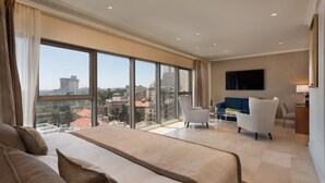 A spacious hotel room with a large bed, a seating area with a blue sofa and chairs, a flat-screen TV mounted on the wall, and a view of the cityscape through large windows. - Herbert Samuel Jerusalem (Jerusalem)