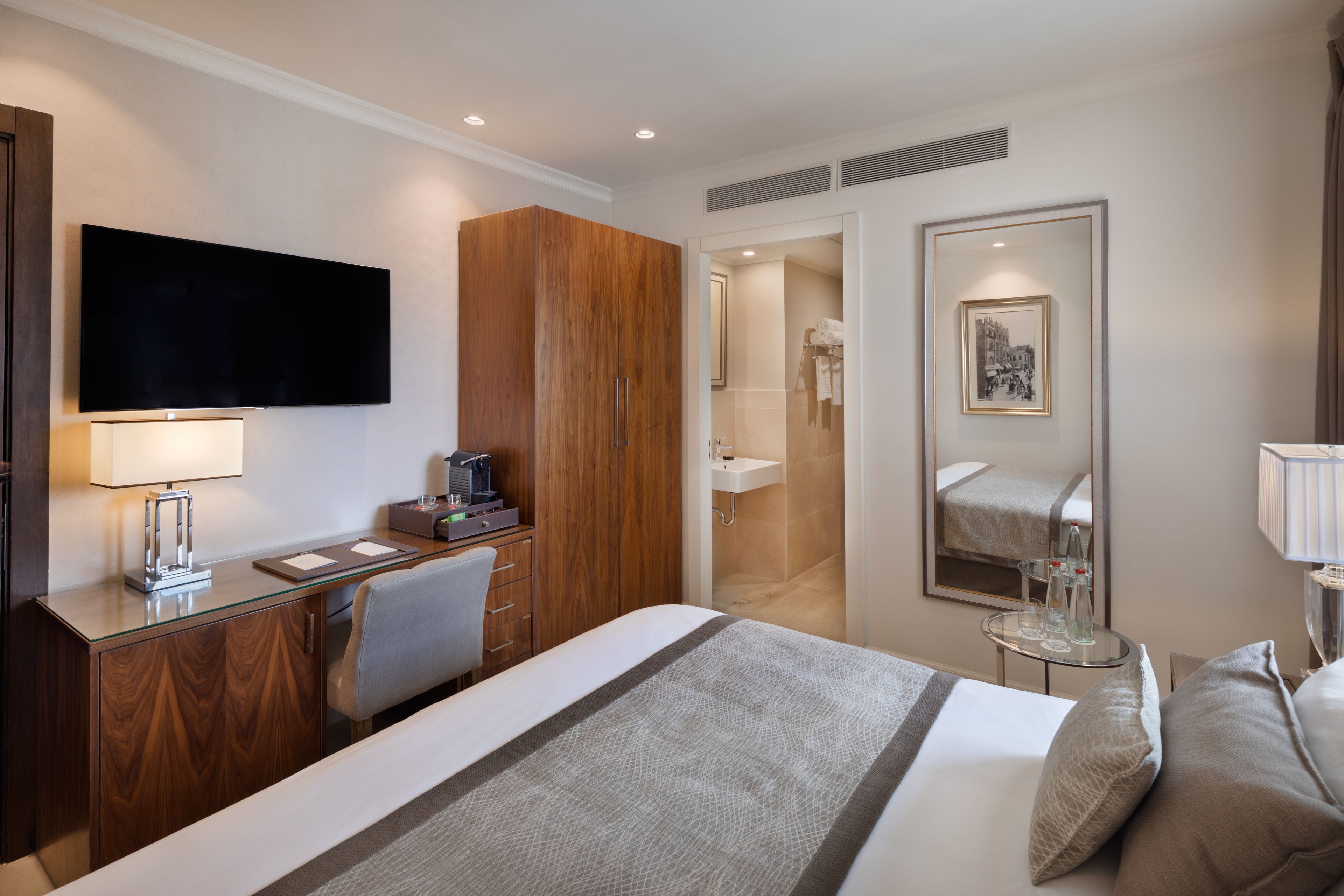 Habitación de hotel con cama, escritorio con lámpara, televisor, espejo y baño visible a través de una puerta abierta.