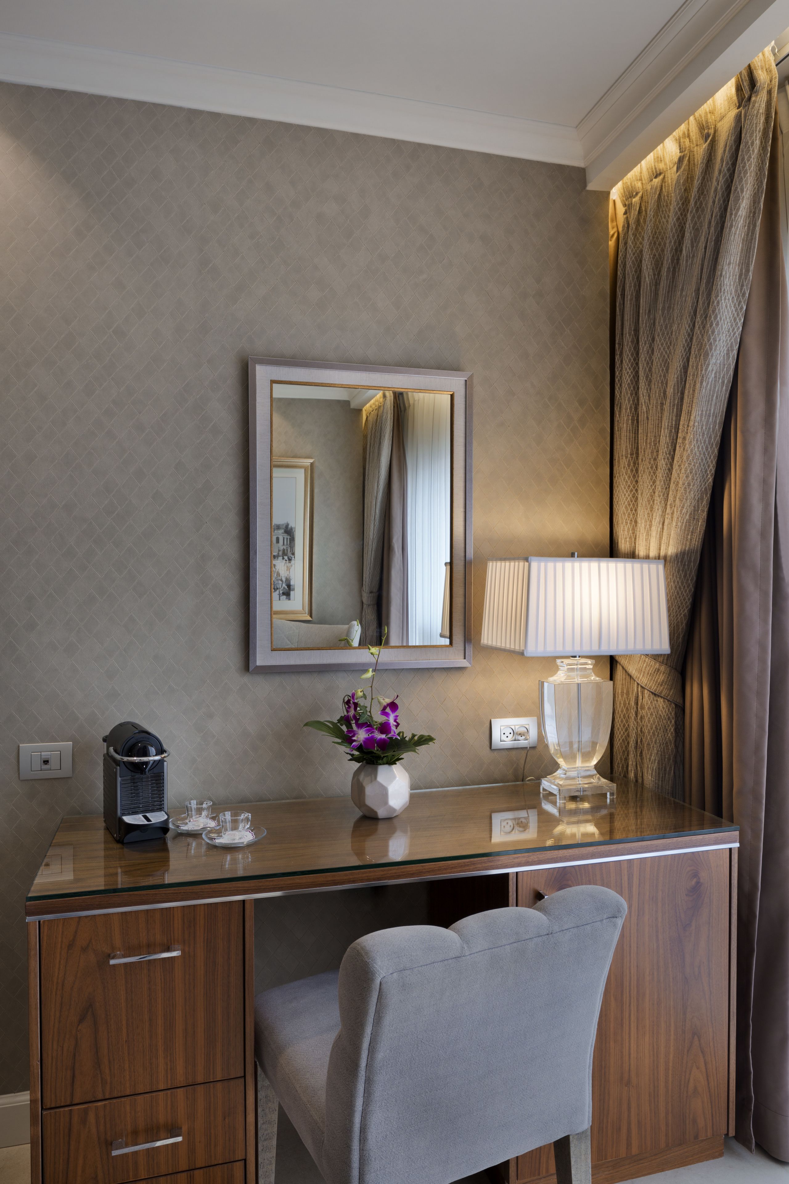 A hotel room with a wooden desk, a chair, a mirror, a lamp, a vase with flowers, and a window with curtains.