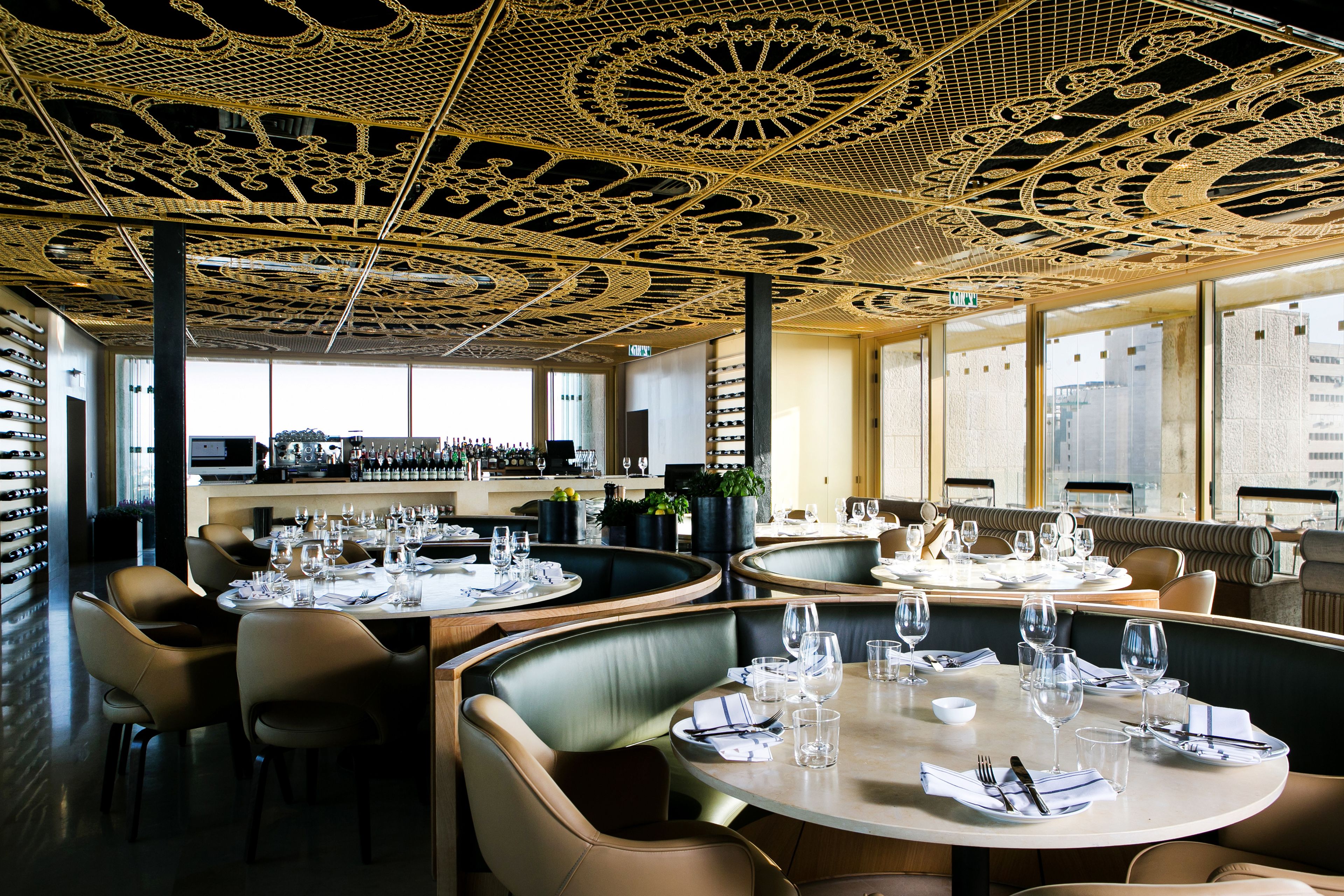 Un restaurante con mesas redondas dispuestas para comer, un techo decorativo y vistas de la ciudad a través de amplias ventanas.