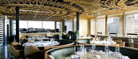 Un ristorante con tavoli rotondi apparecchiati per la cena, un soffitto decorativo e vista sulla città attraverso ampie finestre.