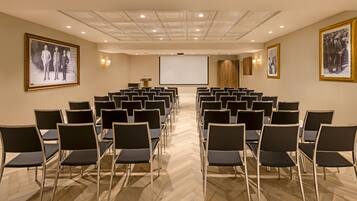 Un salón de conferencias con hileras de sillas, una pantalla de proyector y cuadros en las paredes.