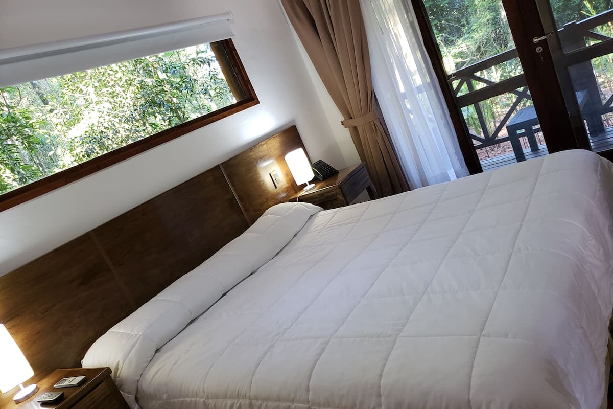 Una cama impecablemente tendida con edredón blanco, cabecera de madera, mesita de noche con lámpara y vista a la vegetación a través de una ventana.