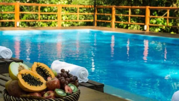Un espace aménagé au bord de la piscine, avec un panier en osier rempli de fruits coupés et de raisins, une serviette enroulée, et une clôture en bois en arrière-plan.