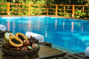 Zona de piscina con una cesta de mimbre llena de fruta cortada y uvas, una toalla enrollada y una valla de madera al fondo.