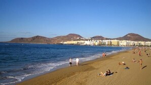 Una playa con gente tomando el sol y nadando, cielo despejado y montañas al fondo.