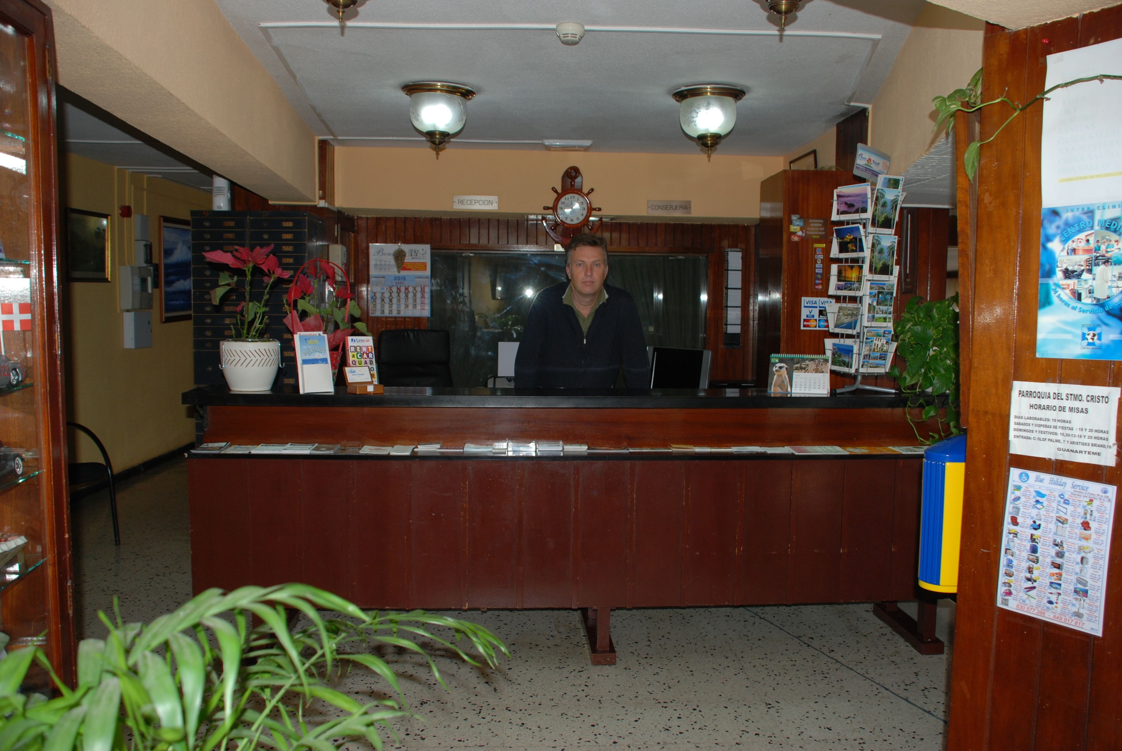 Un mostrador de recepción con un reloj, una planta y un tablón de anuncios con varios avisos.