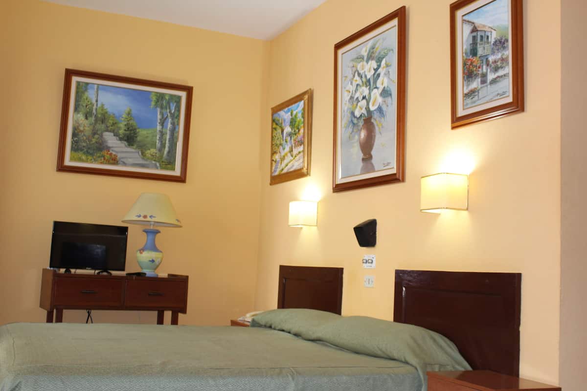 A hotel room with a bed, a desk with a lamp, a television, and three framed paintings on the wall.