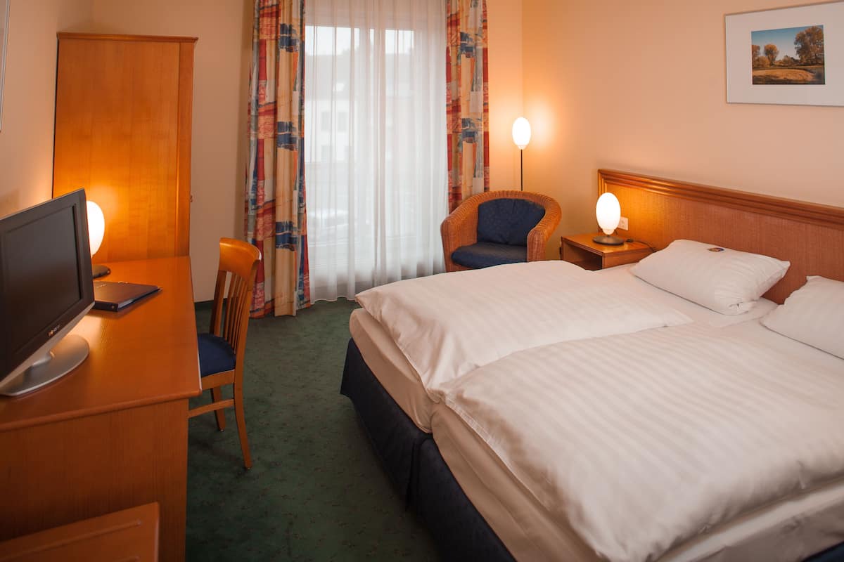 Ein Hotelzimmer mit Bett, Schreibtisch mit Computer, Stuhl, Nachttisch mit Lampe und Fenster mit Vorhängen.