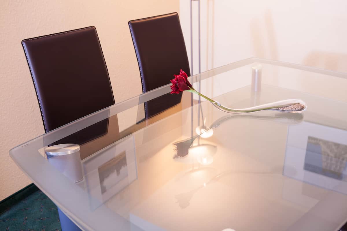 Ein Glastisch mit einer einzelnen roten Rose in einer Vase, zwei schwarzen Stühlen und einem kleinen weißen Gegenstand darauf.