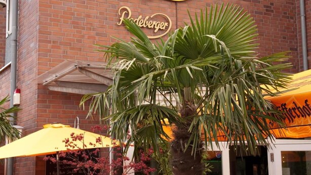 A brick building with a sign reading "XlE BÜDERICH" and "Pöbelinger." There are outdoor tables and chairs, and a palm tree in a planter. - Hotel zum Deutschen Eck (Meerbusch)