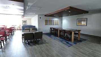 A modern hotel lobby with a long dining table, red and brown chairs, and a blue patterned rug.