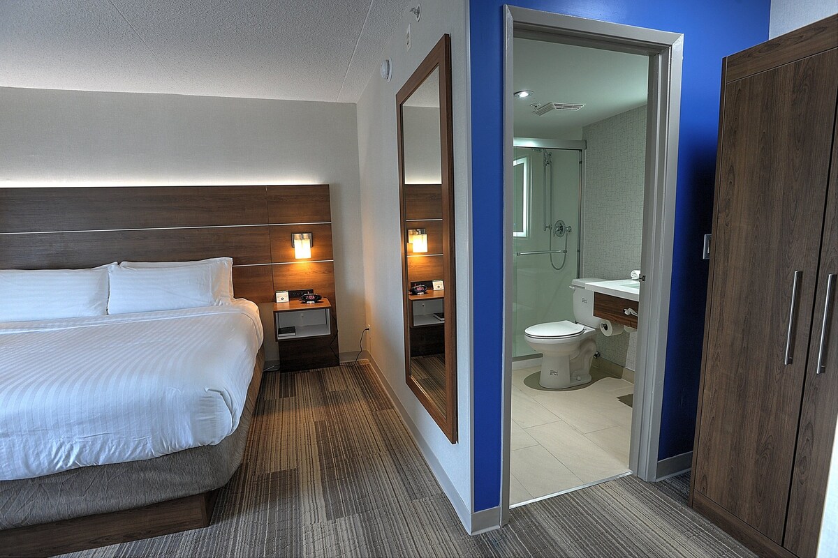 A hotel room with a bed, bedside tables, a mirror, and a bathroom with a shower, toilet, and sink.