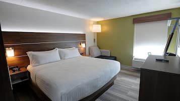 Una cama bien hecha con sábanas blancas, cabecera de madera, lámparas de noche, una mesita de noche con un reloj y un escritorio con un control remoto.