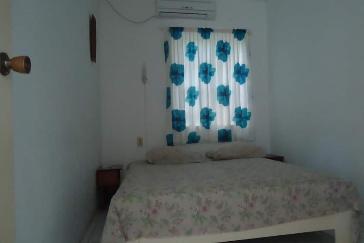 A bedroom with a bed, a nightstand, a wall-mounted air conditioner, and a window with blue floral curtains.