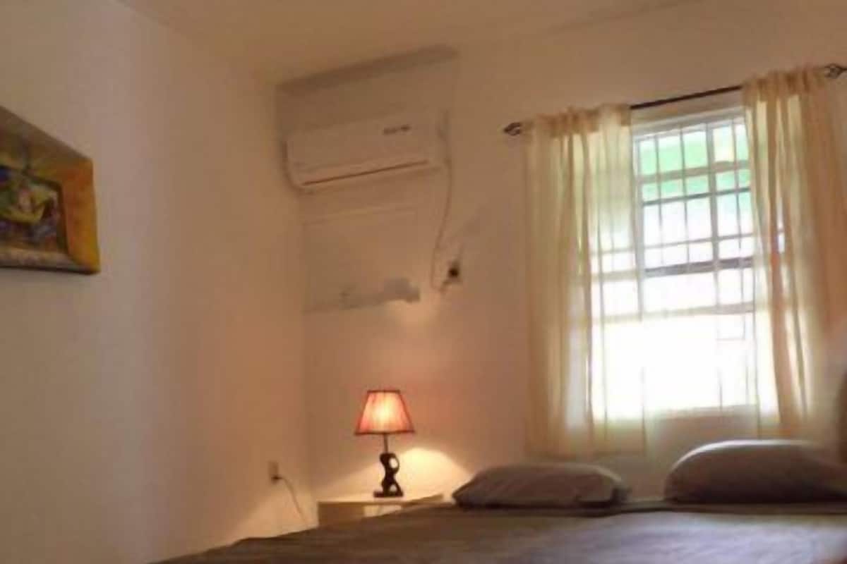 A bedroom with a bed, a nightstand with a lamp, a window with curtains, and an air conditioning unit on the wall.
