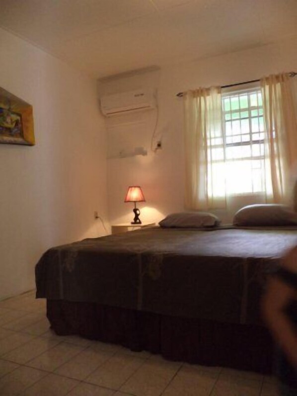 A bedroom with a bed, a nightstand with a lamp, a window with curtains, and an air conditioning unit on the wall.