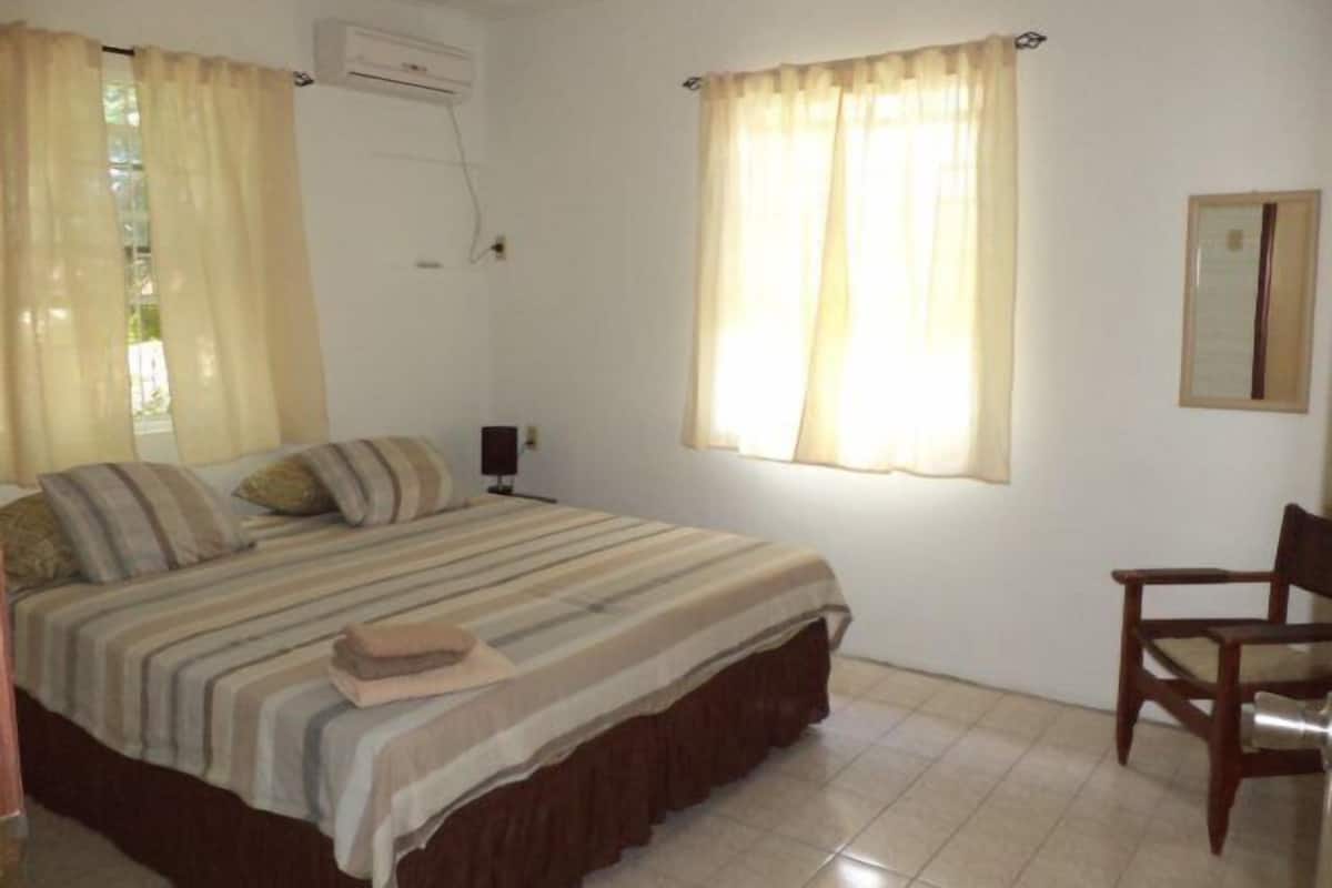 A bedroom with a bed, a chair, a nightstand, an air conditioning unit, and a window with curtains.