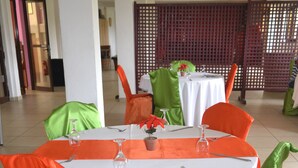 Un espace repas avec des tables rondes, des housses de chaise orange et verte, et des nappes blanches.