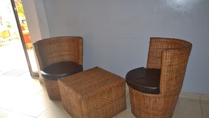 Deux fauteuils en osier avec des coussins noirs et un pouf assorti, dans une pièce dont le sol est recouvert de carreaux et dont un des murs est uni.