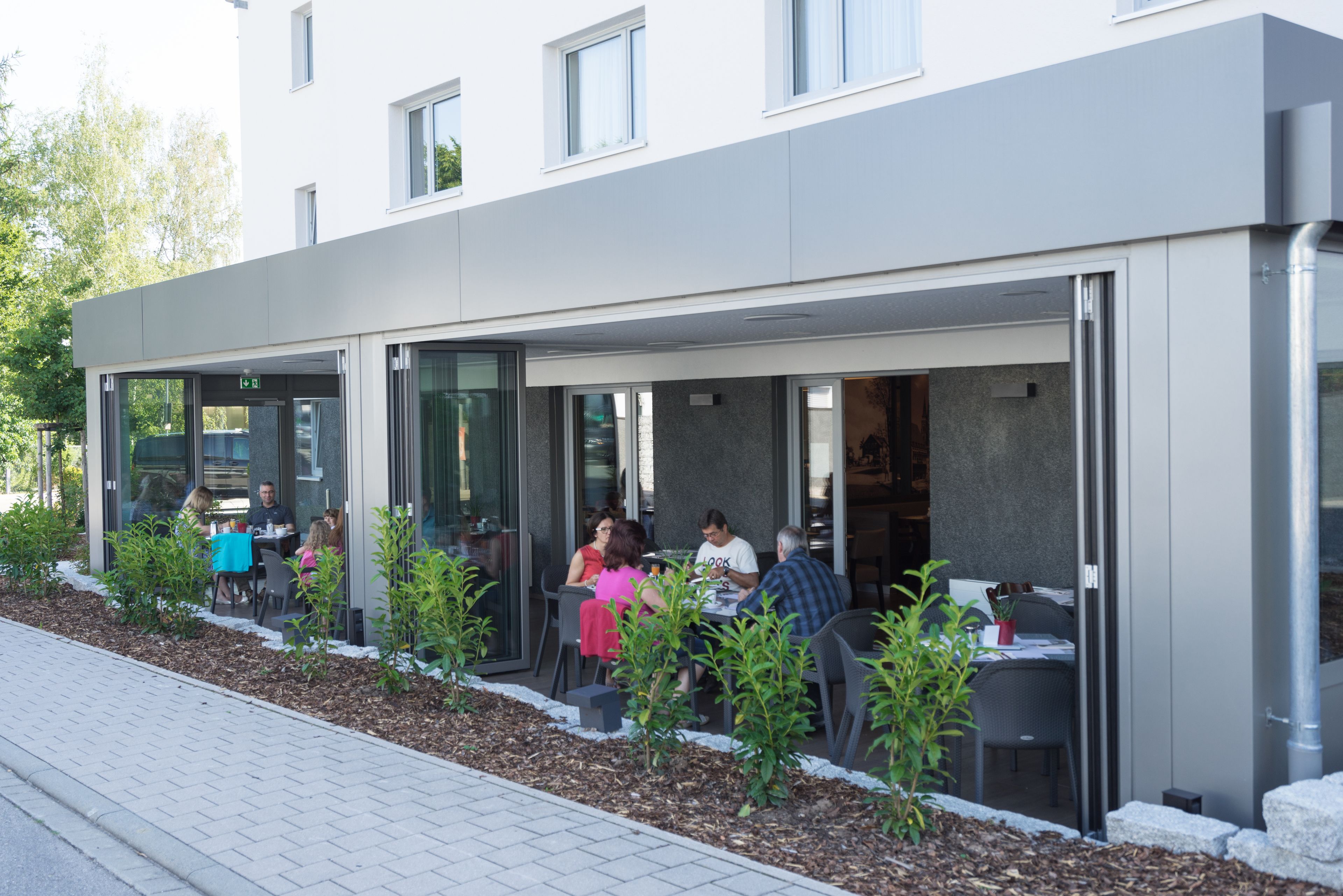 An outdoor seating area with tables and chairs, surrounded by small plants and a pathway.