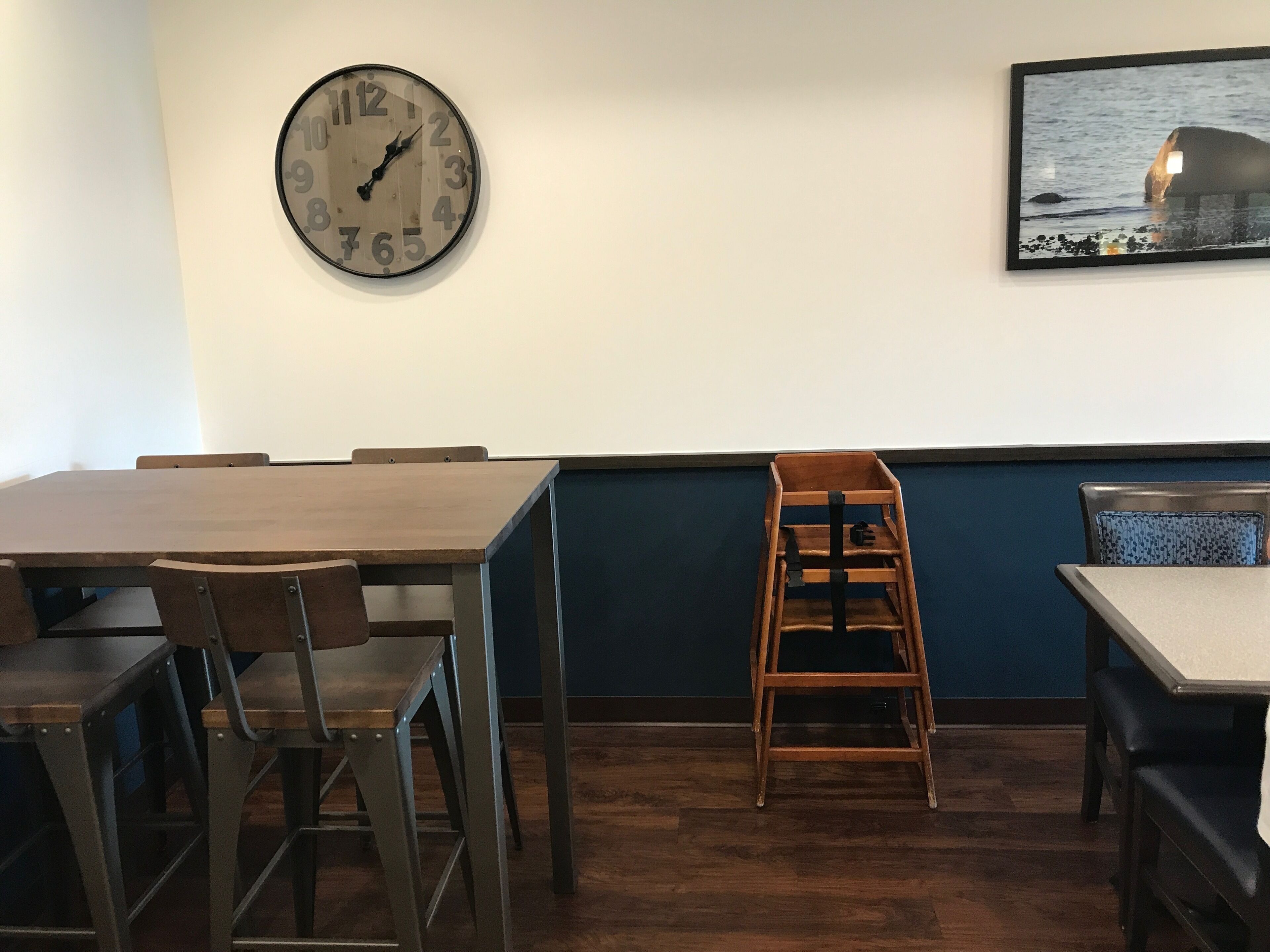 A restaurant interior with a wooden table and chairs, a clock on the wall, and a painting of a seascape.