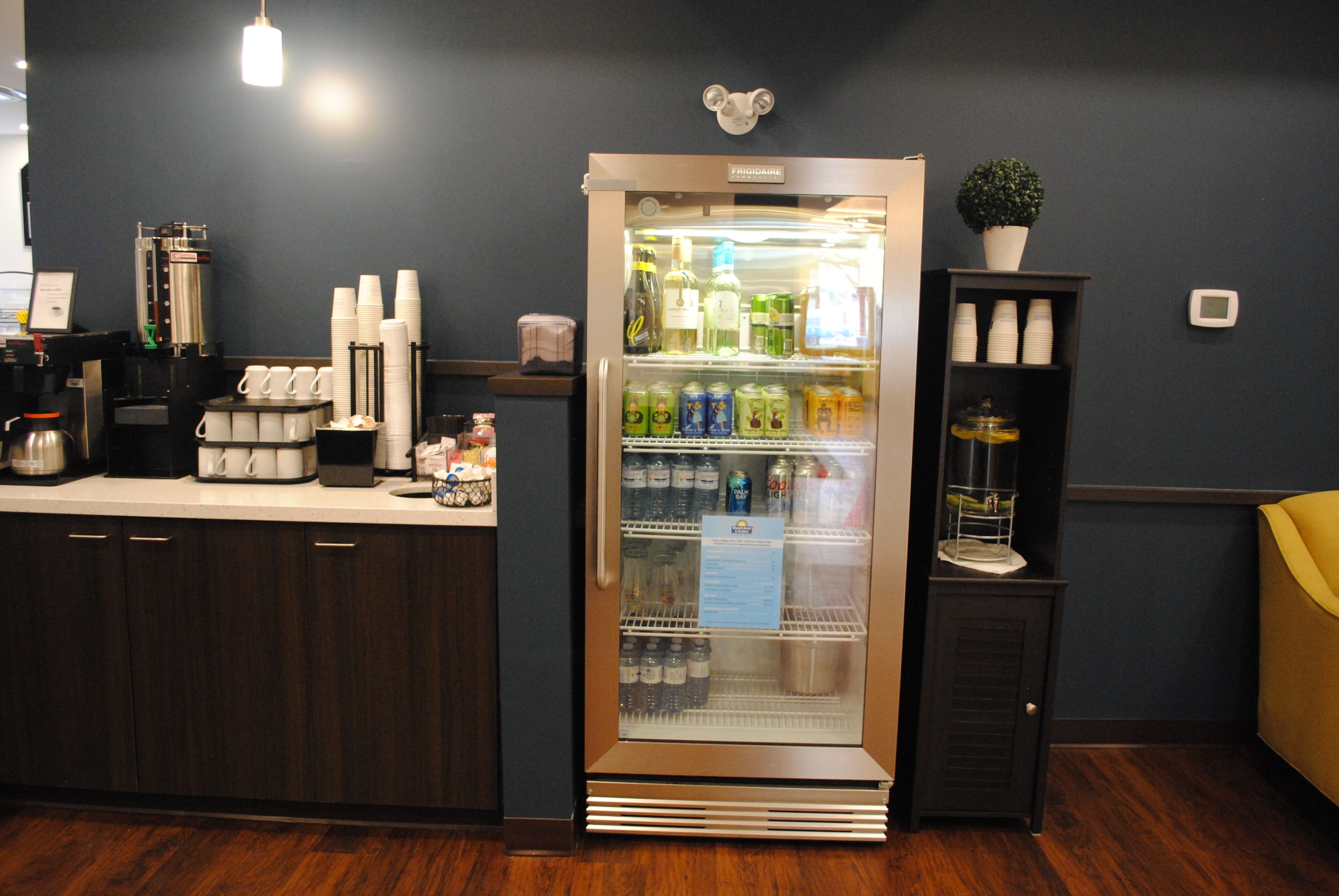A kitchen area with a refrigerator stocked with beverages, a coffee machine, and a shelf with cups.
