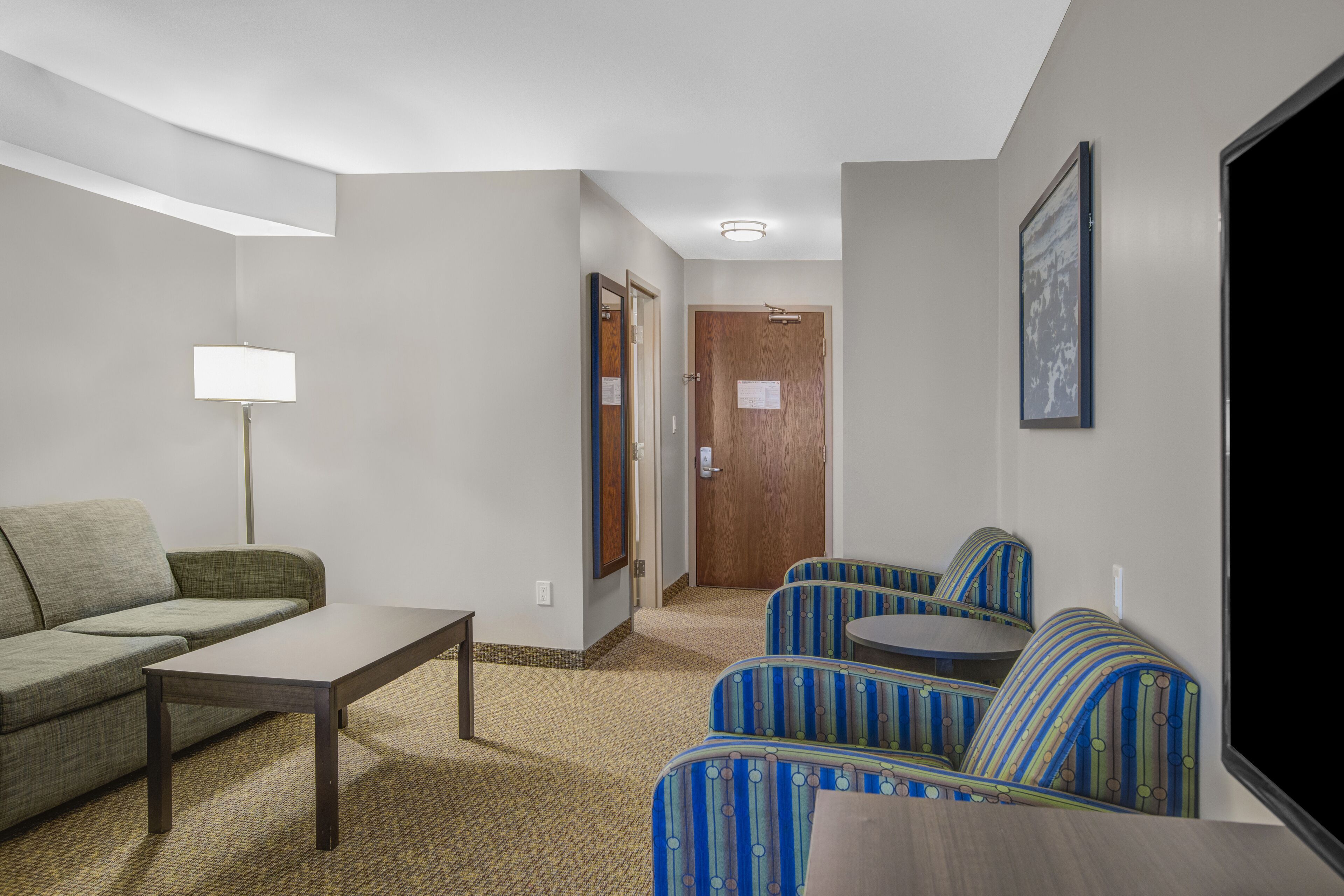 A hotel room with a sofa, a small table, a lamp, and a seating area with patterned chairs.