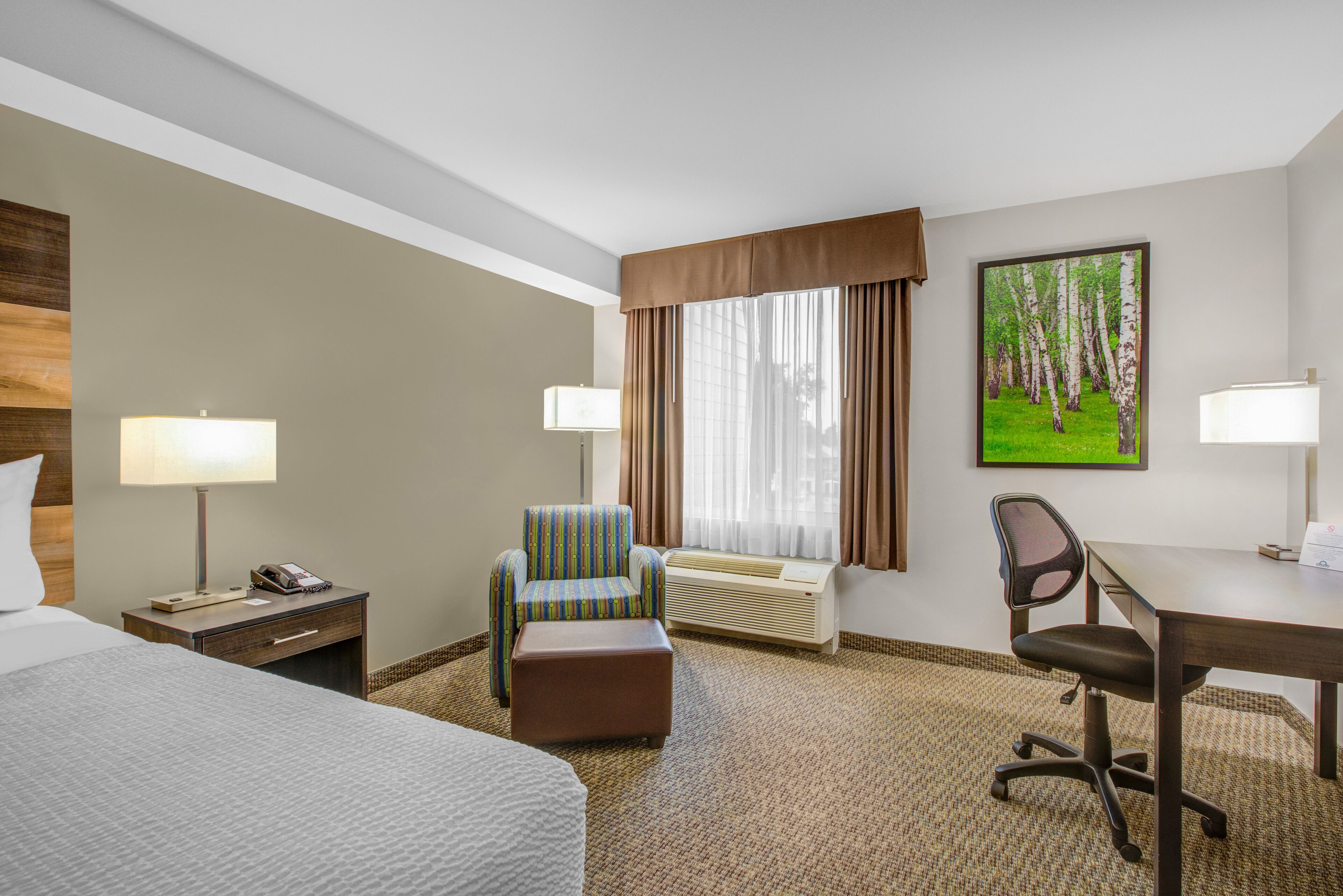 A hotel room with a bed, a desk, a chair, an ottoman, a lamp, a telephone, and a large window with curtains.