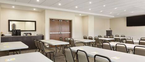 A conference room with multiple tables and chairs, a kitchenette, a large screen, and a door with a mirror.