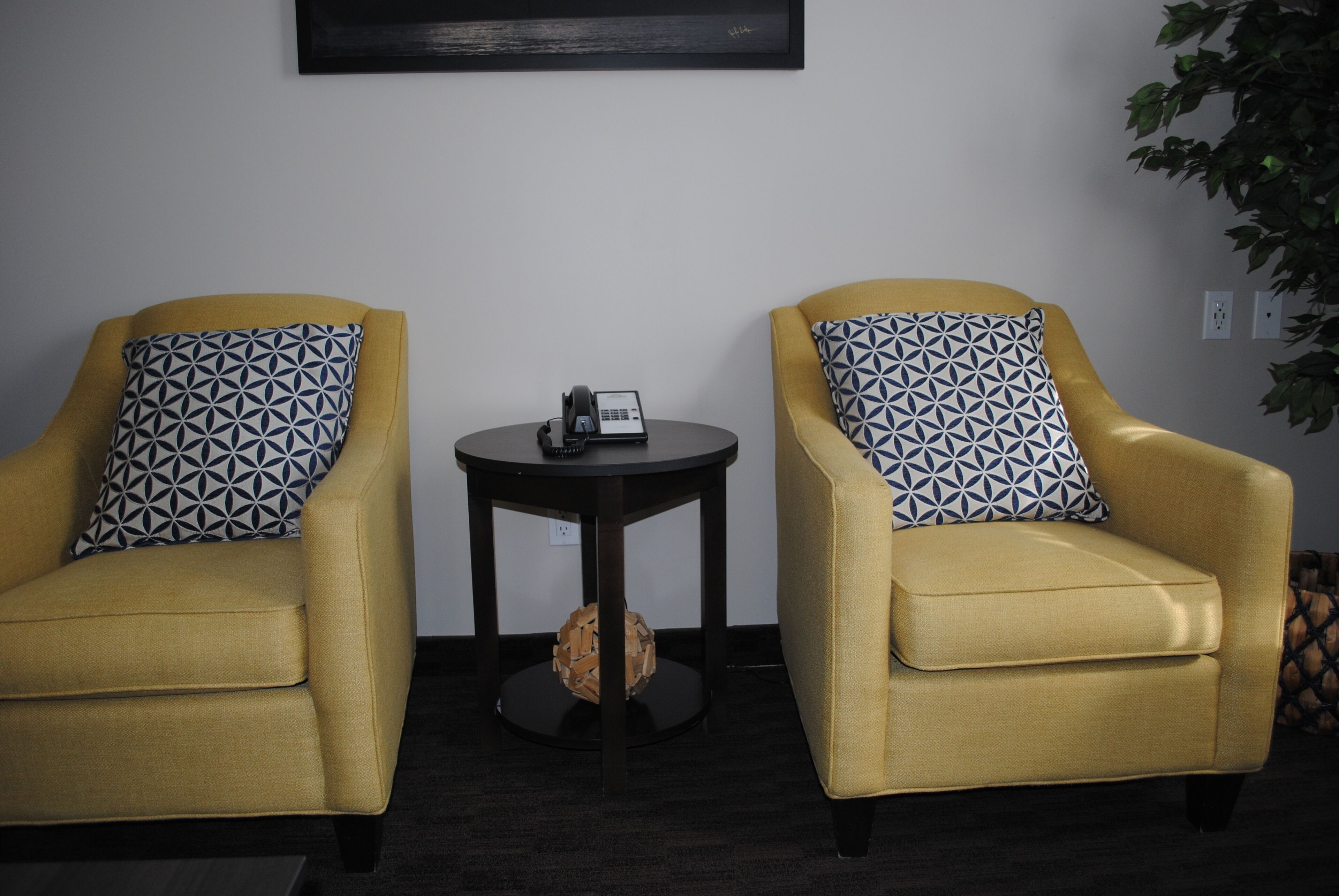 Two yellow armchairs with geometric patterned pillows, a small round table with a telephone, and a wall-mounted artwork.