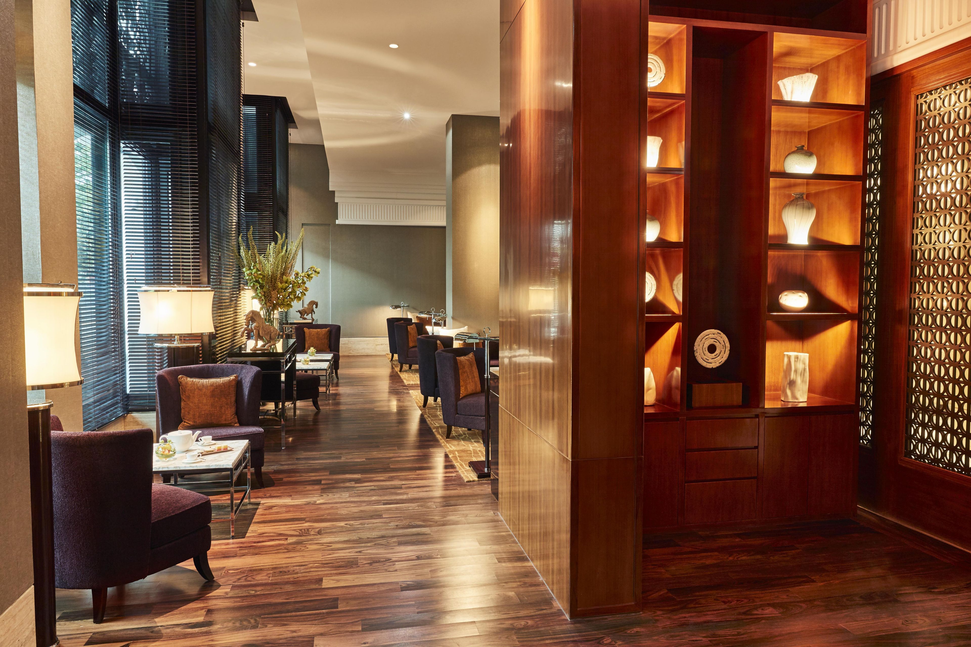 Vestíbulo de hotel con piso de madera, zona de estar con sillones, mostrador de recepción y una pared decorativa con estanterías.