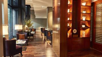 Vestíbulo de hotel con piso de madera, zona de estar con sillones, mostrador de recepción y una pared decorativa con estanterías.