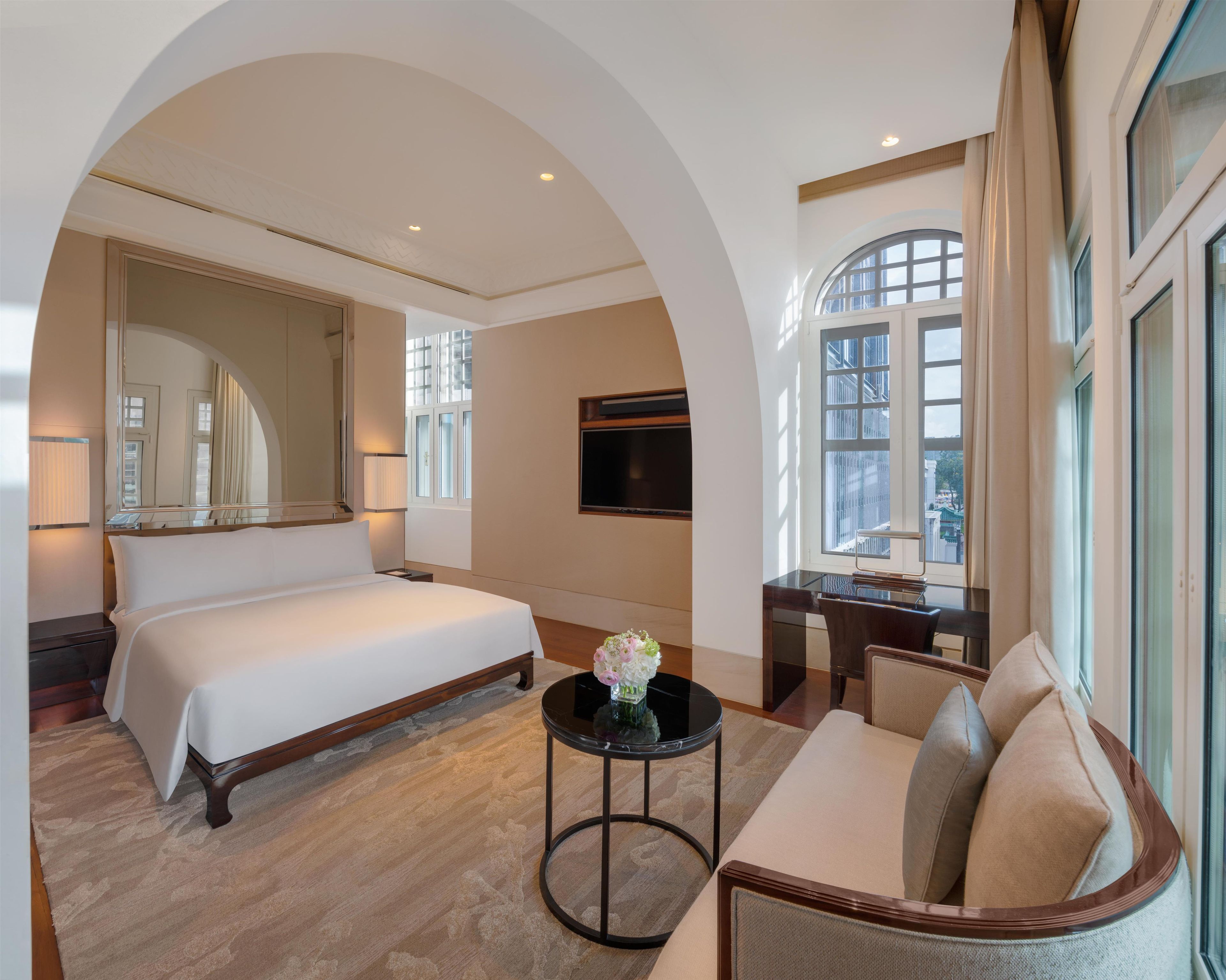 Habitación de hotel con una cama grande, un escritorio, una silla, un televisor y vistas a la ciudad desde las ventanas.