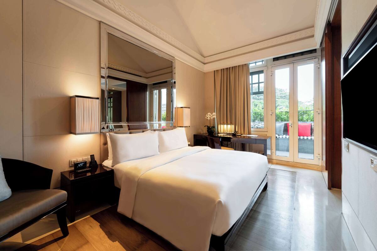 A hotel room with a large bed, a bedside table, a nightstand, a desk, a chair, a large mirror, a TV, and a balcony with a view.