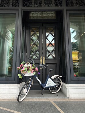Uma bicicleta com um cesto de flores estacionada em frente a uma porta preta com painéis de vidro decorativos.