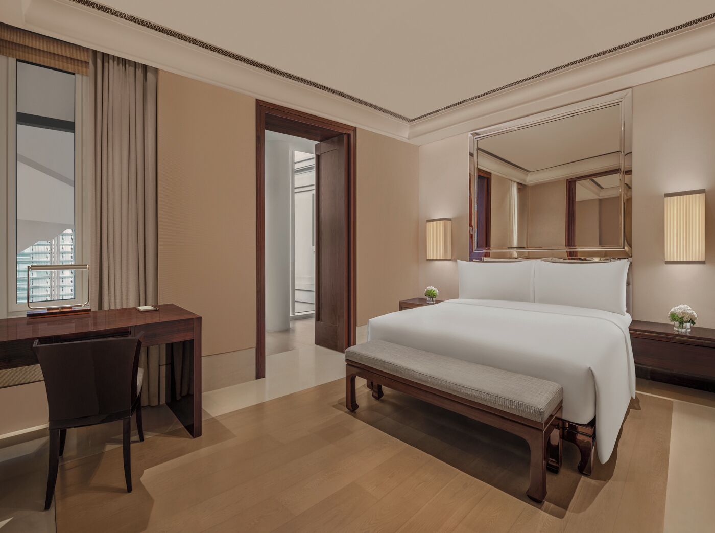 Habitación de hotel con una cama grande, un escritorio, una silla, un banco, un espejo y una ventana con cortinas.