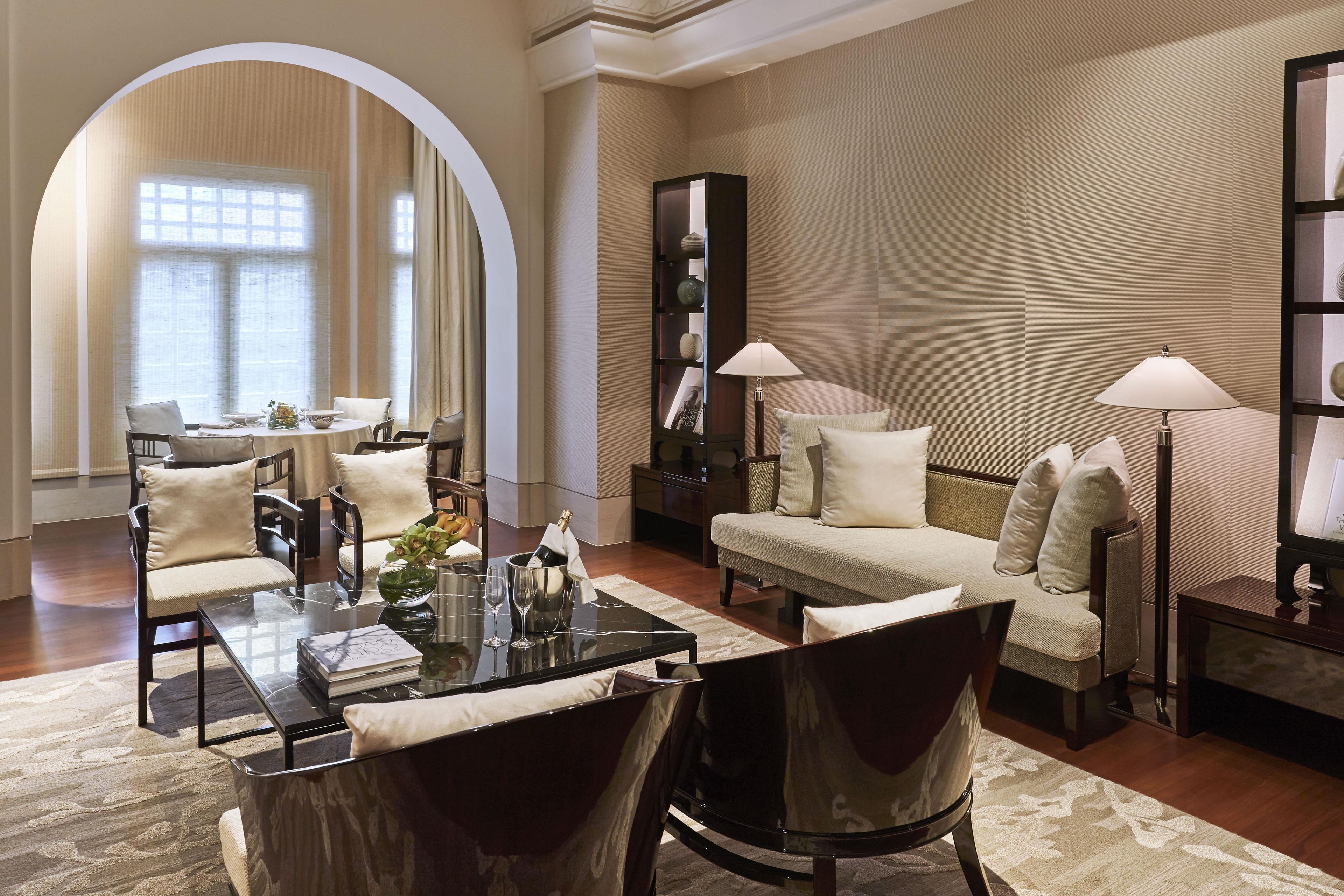 A well-lit living room with a glass coffee table, two armchairs, a sofa, and a bookshelf.