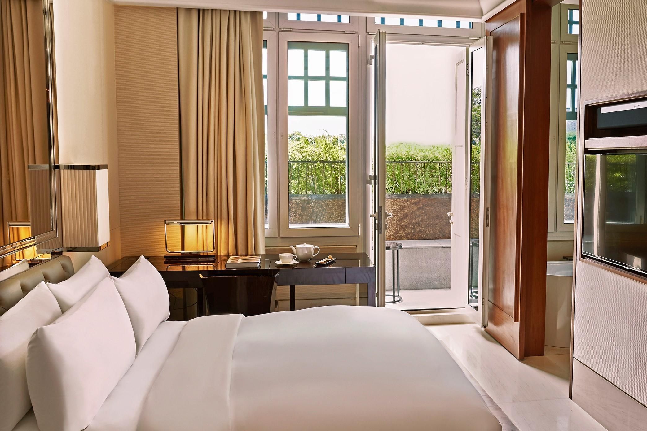 A hotel room with a large bed, a desk with a chair, a television, and a view of greenery outside.