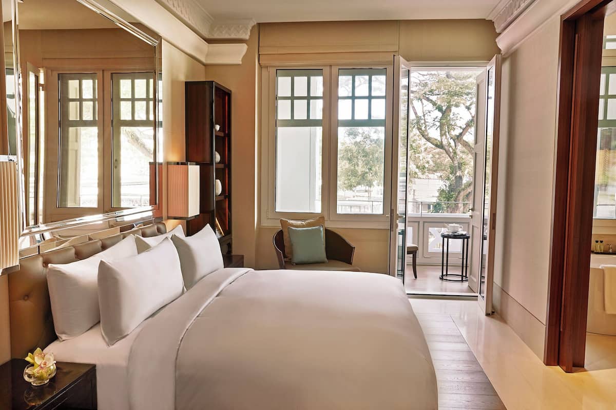 A spacious bedroom with a large bed, a nightstand, a bookshelf, and a view of the outdoors through a large window.