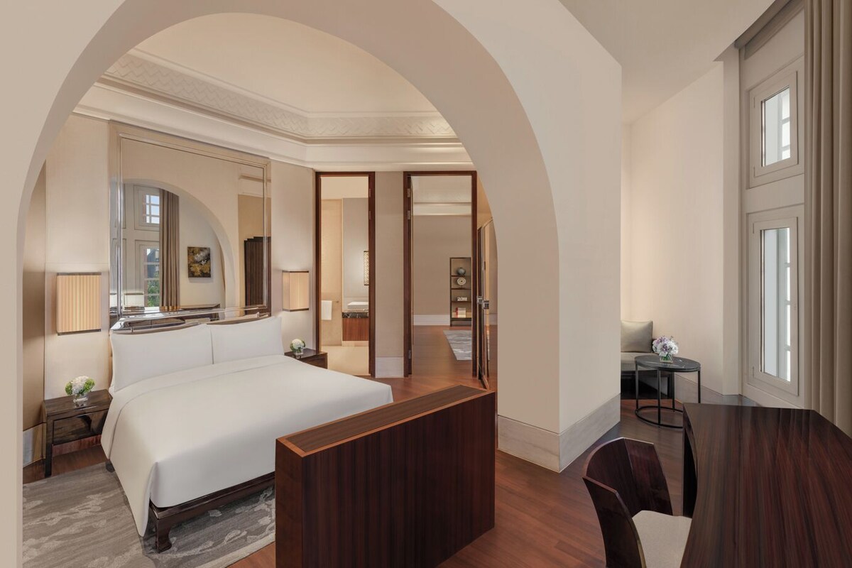 A hotel room with a large bed, a wooden desk, a chair, a mirror, and a view of the outside through a window.