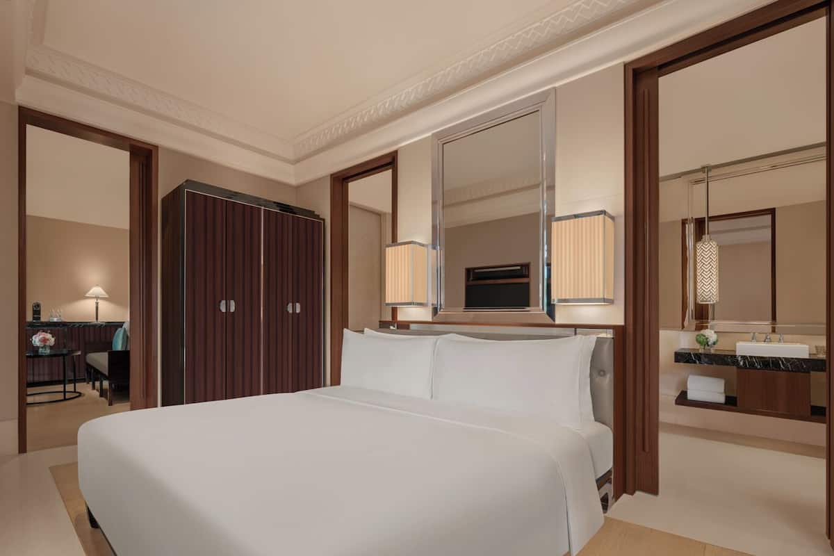 A hotel room with a large bed, a desk with a lamp, a wardrobe, and a bathroom with a mirror and sink.
