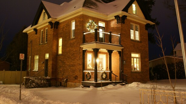 A brick house with a balcony, snow on the roof, and a lit front porch. - Danby House (Grey Highlands)
