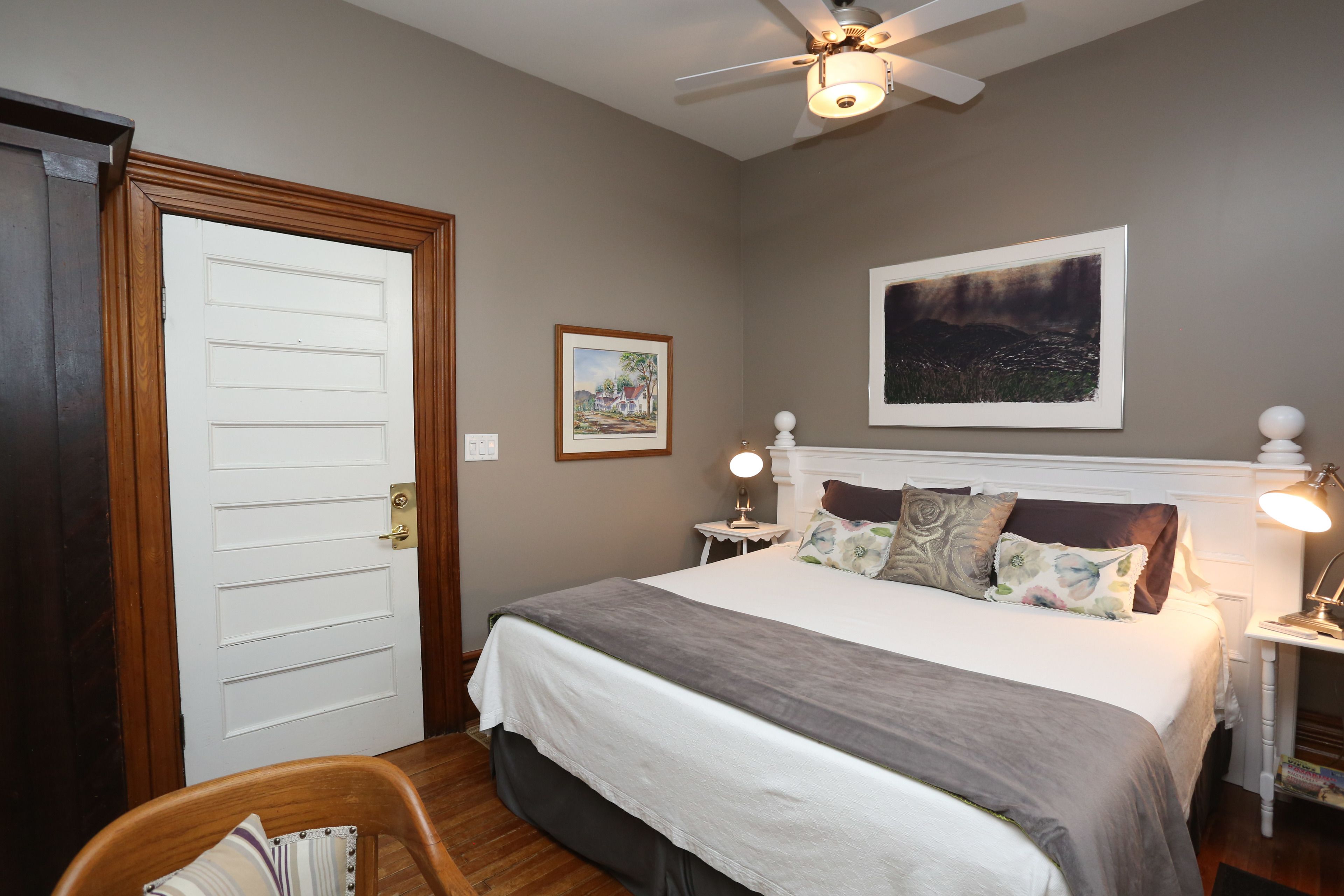A bedroom with a bed, a ceiling fan, a nightstand with a lamp, a framed picture on the wall, and a door.