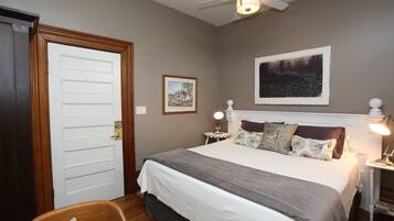 A bedroom with a bed, a ceiling fan, a nightstand with a lamp, a framed picture on the wall, and a door.