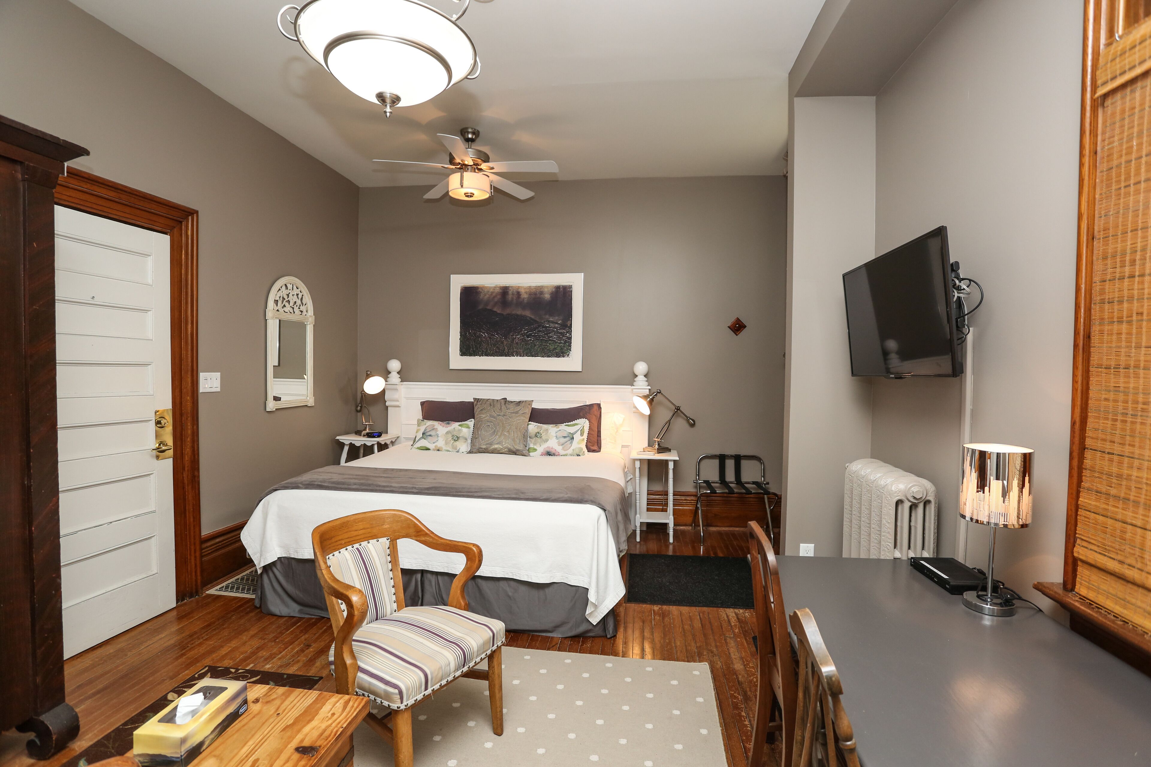 A bedroom with a bed, a chair, a desk, a lamp, a ceiling fan, a TV, and a radiator.
