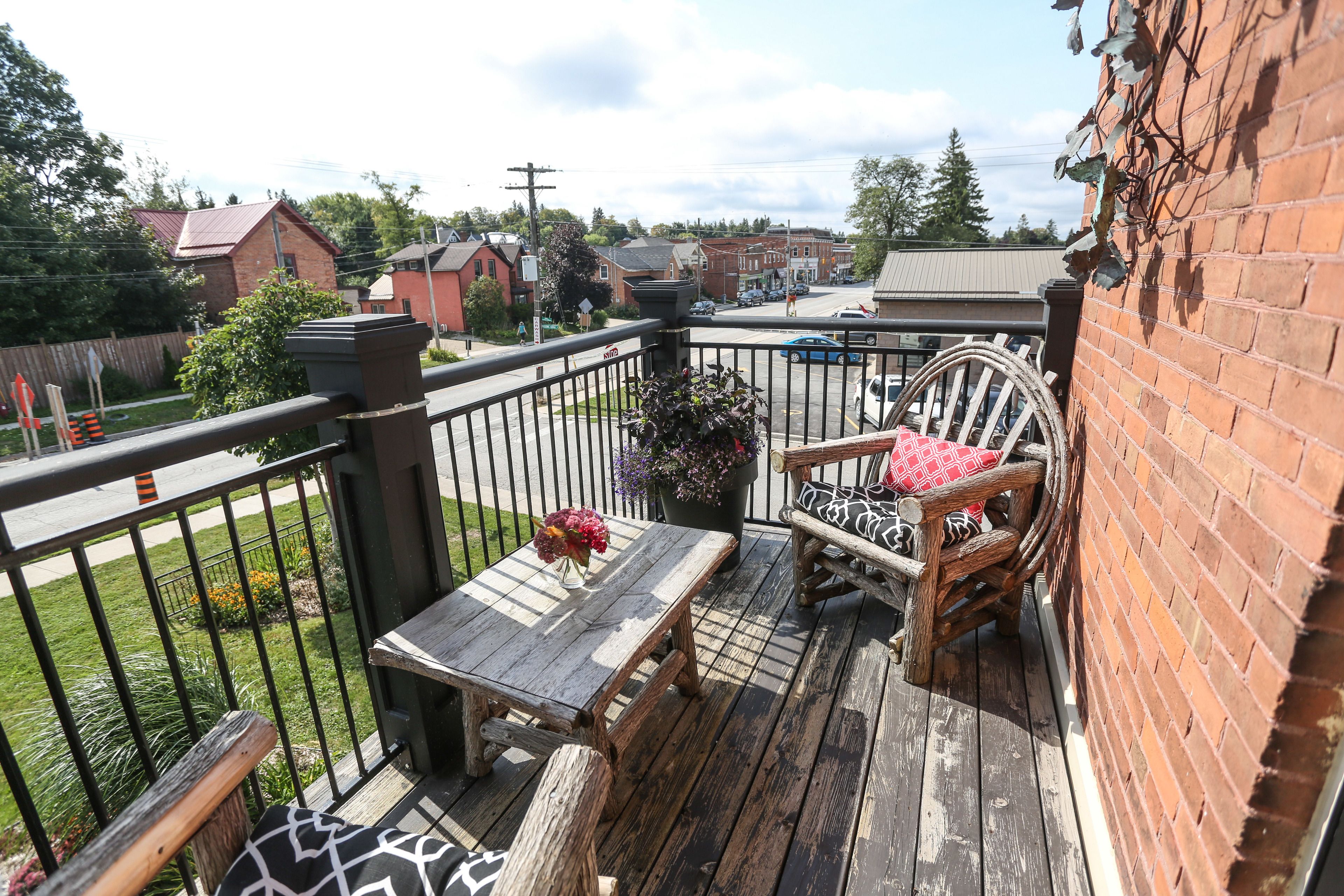 A wooden deck with a table and chairs, a brick wall, and a view of a street with buildings and a grassy area.
