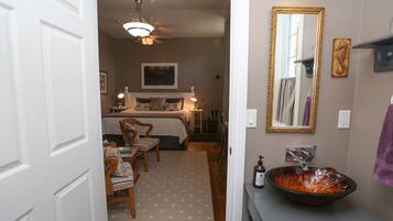 A bedroom with a bed, bedside tables, a chair, a desk, and a bathroom with a sink and mirror.