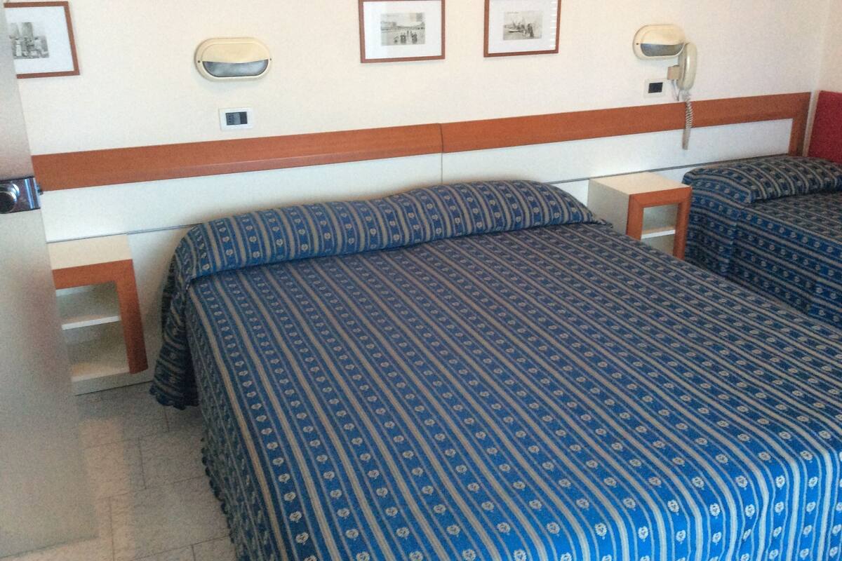 A hotel room with two beds, each with a patterned blue bedspread. There are framed pictures on the wall, a bedside telephone, and a small table between the beds.